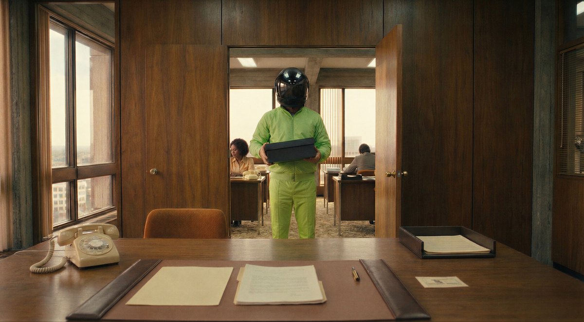 A courier in a neon green tracksuit and helmet stands in the office doorway, holding a black box